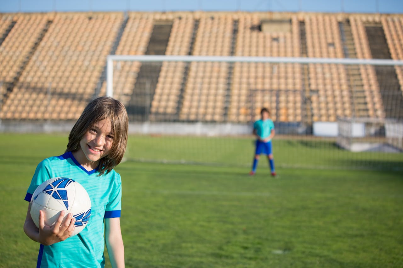 Two kids enjoying a game of soccer at an outdoor field with a stadium backdrop.